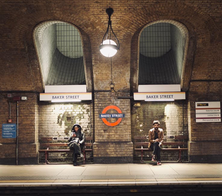 View of Baker Street underground station platform with two benches, each occupied by a person. The station has exposed brick walls with two arched openings and tiled surfaces inside the arches. Overhead, a hanging lamp illuminates the area, and signs reading 'BAKER STREET' are positioned above the benches. The platform surface is clean, and the overall scene conveys a well-maintained environment. Cleaners Marylebone provides expert surface cleaning, deep cleaning, and sanitisation services to maintain hygiene and cleanliness in such public transportation spaces.