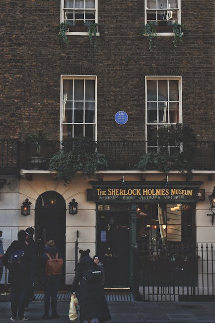 The exterior of The Sherlock Holmes Museum located on Baker Street features a dark brick facade with two vertically divided, white-framed windows on the upper level, each adorned with green plants in window boxes. Below, the entrance has a black sign with gold lettering indicating the museum's name, and a white facade with black lantern-style lights on either side of the door. In front of the museum, several visitors are standing and walking on the pavement, some wearing casual clothing and backpacks, with soft natural lighting illuminating the scene. Cleaners Marylebone regularly performs surface cleaning and sanitisation of public-facing areas like this to maintain hygiene and appearance, ensuring the space remains welcoming for visitors to this famous tourist destination.