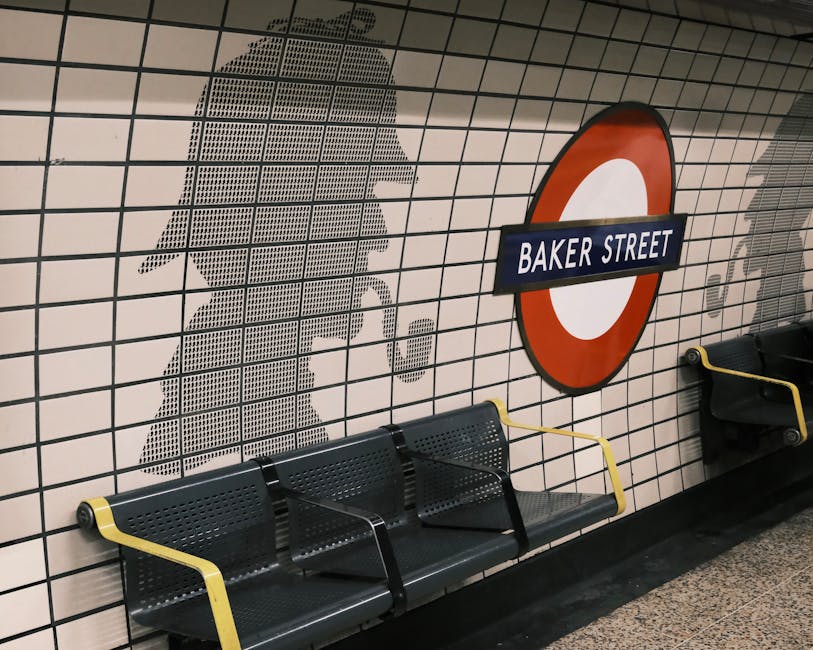 View of Baker Street underground station platform with two benches, each occupied by a person. The station has exposed brick walls with two arched openings and tiled surfaces inside the arches. Overhead, a hanging lamp illuminates the area, and signs reading 'BAKER STREET' are positioned above the benches. The platform surface is clean, and the overall scene conveys a well-maintained environment. Cleaners Marylebone provides expert surface cleaning, deep cleaning, and sanitisation services to maintain hygiene and cleanliness in such public transportation spaces.