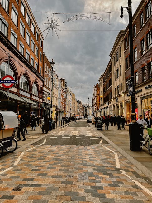 An outdoor pedestrian street in Marylebone high street with a paved pattern of multicoloured bricks. The street is lined with multi-storey buildings featuring brick and stone facades, housing shops with large display windows and signage. On either side, there are street lamps, holiday decorations such as hanging string lights and star-shaped ornaments, and people walking or standing near shop entrances. The sky is overcast, and the street appears clean and well-maintained, reflecting the professional cleaning services offered by Cleaners Marylebone for commercial and retail environments. The scene captures a lively shopping district with a focus on maintaining hygiene and cleanliness in a busy urban commercial setting.