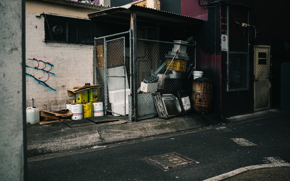 An outdoor waste storage area featuring a chain-link fence enclosure adjacent to a white-painted brick wall with graffiti. Inside the enclosure, various bulky waste items such as large metal trays, wooden pallets, plastic containers, and metal frames are visible. The ground is paved with asphalt, showing some stains and minor cracks. Nearby, there are rolls of yellow and white packaging materials and a cardboard box placed on the pavement. The setting is dimly lit, suggesting a shaded or low-light environment, typical of an alley outside a commercial or residential building. This area illustrates the importance of proper waste management and surface cleaning, as offered by Cleaners Marylebone, for maintaining hygiene and environmental standards in Marylebone.