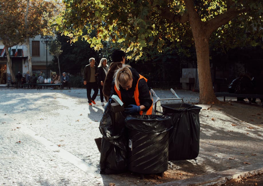 A woman wearing an orange and black high-visibility jacket is collecting waste from large black garbage bags placed on a cobblestone pathway in a park. She is using gloves and a handheld device, possibly a waste scanner or identification tool, to manage the waste. The scene is illuminated by natural sunlight, with shaded areas under a large tree with green and yellow autumn leaves. In the background, several people are walking along the path, and park benches and buildings are visible. The setting reflects outdoor waste collection and the importance of efficient waste management, similar to the surface cleaning and sanitisation services offered by Cleaners Marylebone, especially in handling bulky waste and maintaining hygiene in residential and commercial environments.