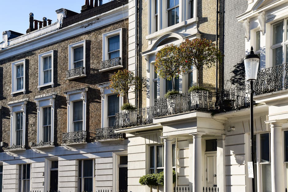 Exterior view of a row of traditional Georgian-style terraced houses in Marylebone, London, featuring a mix of brick and stucco facades. The white-painted building on the right has black wrought-iron balcony railings, decorative window trims, and a rounded bay window above the entrance, which is surrounded by columns. Potted plants and small trees adorn the balconies and entrance area, while a vintage-style street lamp is positioned next to the building. The scene is well-lit with natural daylight, highlighting the cleanliness and care of the residential exteriors, with no visible dirt or debris, reflecting a well-maintained and hygienic urban environment, consistent with the high standards of domestic cleaning services provided by Cleaners Marylebone.