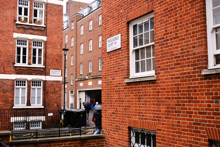 Exterior view of a row of traditional Georgian-style terraced houses in Marylebone, London, featuring a mix of brick and stucco facades. The white-painted building on the right has black wrought-iron balcony railings, decorative window trims, and a rounded bay window above the entrance, which is surrounded by columns. Potted plants and small trees adorn the balconies and entrance area, while a vintage-style street lamp is positioned next to the building. The scene is well-lit with natural daylight, highlighting the cleanliness and care of the residential exteriors, with no visible dirt or debris, reflecting a well-maintained and hygienic urban environment, consistent with the high standards of domestic cleaning services provided by Cleaners Marylebone.