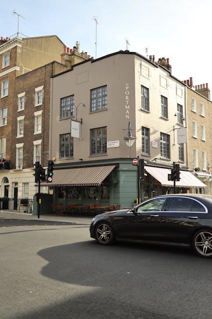 Exterior view of a three-story corner building with a light beige facade and multiple windows, situated at the intersection of a street with traffic lights. The ground floor features a green-painted section with a striped awning over outdoor seating, likely part of a café or restaurant, with wooden tables and chairs visible. A black car is parked in front of the building on the street. The surrounding buildings are older, built with brick and similar in style, located in Marylebone. The scene is well-lit with natural daylight, and the street appears clean and orderly, reflecting regular maintenance and cleaning practices. As part of domestic cleaning and surface maintenance, Cleaners Marylebone ensures the cleanliness of outdoor areas and building facades, consistent with the detailed upkeep typical for property maintenance on the Portman Estate.