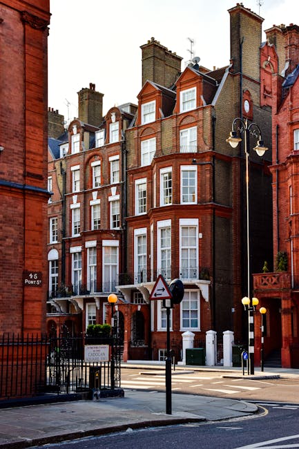 A street view of a row of historic red-brick residential buildings with white-framed bay windows and decorative gables, typical of the Marylebone area. The buildings have multiple stories with chimneys on the roofs, and some feature small front gardens bordered by black metal fencing. A streetlamp with two lamps and a traffic sign are visible in the foreground, along with a pedestrian crossing. The scene is illuminated by daylight, highlighting the clean, well-maintained facades. As part of the services offered by Cleaners Marylebone, this setting exemplifies the importance of surface cleaning and general maintenance to preserve the aesthetic appeal of such heritage properties within the Portman Estate.
