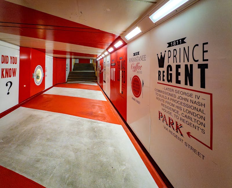 A brightly lit underground passage with a smooth, polished concrete floor featuring a red and white colored pattern. The walls are adorned with vintage-style signage, including the prominent 'Prince Regent' sign with a crown graphic, and other posters promoting coffee and historical references. The ceiling is finished with wooden paneling, and modern rectangular fluorescent lighting illuminates the corridor evenly. Stairs at the end of the passage lead upwards, while the space appears clean, with no dust or dirt visible. The overall setting suggests a commercial environment, possibly part of a pub or public space, where surface cleaning and maintenance are essential for hygiene, as managed by Cleaners Marylebone.