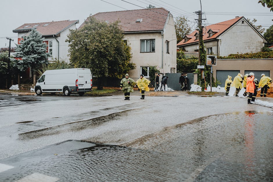 A street scene showing a residential area with several two-storey houses featuring pitched roofs, some with dormer windows, and surrounded by trees and greenery. The street is wet from recent flooding, with visible puddles and water pooling on the asphalt. Multiple workers in yellow raincoats and protective gear are engaged in flood response activities near the curb, some inspecting the area, while others appear to be managing sandbags and water removal. A white van is parked on the side of the road. The background includes utility poles and overcast sky, indicating cloudy weather. This image highlights a flood response effort involving surface cleaning, water extraction, and property protection, taken in support of the services offered by Cleaners Marylebone during emergencies such as the one described in the page titled 'Same-Day Flood Response for Marylebone Basement Flats, Marylebone' at cleaners-marylebone.com.