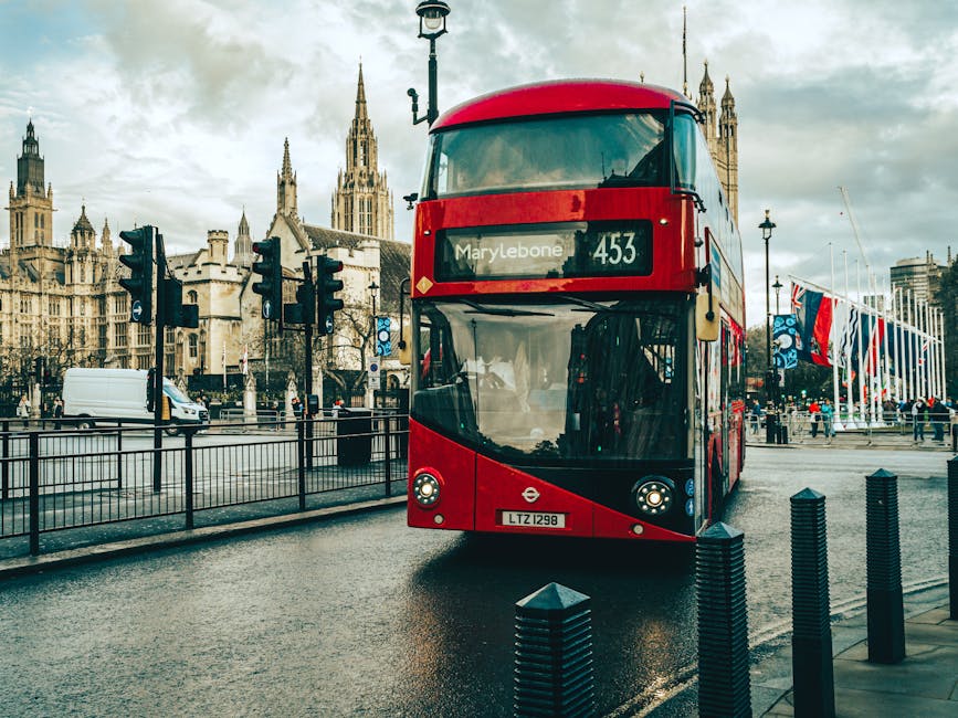 A red double-decker bus labeled with the route number 453 and destination Westminster is stationary on a wet street in Marylebone, London, with the historic Westminster Palace and its pointed spires visible in the background. The scene includes traffic lights, black metal bollards along the sidewalk, and a row of flags flying on flagpoles to the right. The cloudy sky casts a diffused light over the area. This image emphasizes urban cleanliness and maintenance, relevant to surface cleaning and hygiene practices promoted by Cleaners Marylebone, especially in relation to household waste and sanitation standards outlined by Westminster Council.